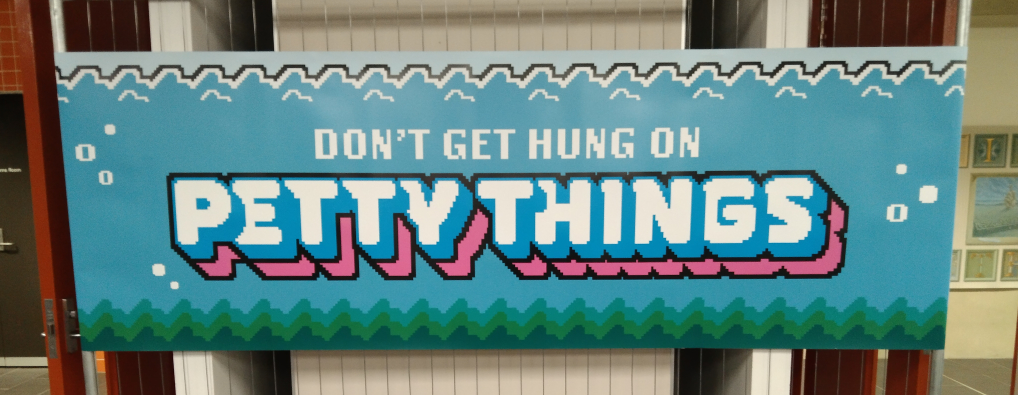 The mural reads 'Don't get hung up on petty things'. The letters are white in a heavy weight uppercase pixel font. Don't Get Hung Up On is a geometric sans serif and Petty Things is a bubble font. Petty things has a blue 3D applied to it and a black outline, with a pink drop shadow with a black outline. There are bubbles on either side of the text, with layered seaweed or moss along the bottom and blue, white, and black waves along the top with a small bit of sky behind it.