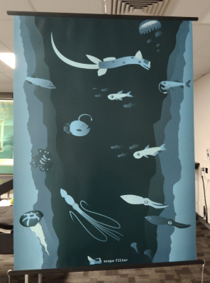 The poster is blue and in a vertical orientation hanging from the ceiling. It features cliff walls on the left and right sides and deep sea creatures in a flat vector style on the front. The logo for scope filter is at the bottom of the poster.