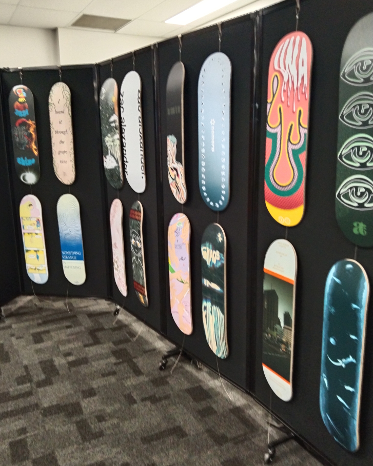 Skateboard decks minus the wheels are hanging against a black wall held up by wire. Designs are printed as a sticker which is stuck on the board.