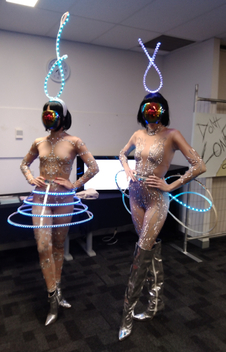Two ladies are wearing a science fiction outfit. They are wearing a flesh tone outfit with silver beads on them, black bob cut wigs, and skirts and wings made of LED rings. Both are wearing twisted lights on their head.